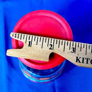 May include: A red lid covers a blue and orange container. A wooden ruler is placed across the lid, measuring approximately 3 inches. The container sits against a bright blue background.