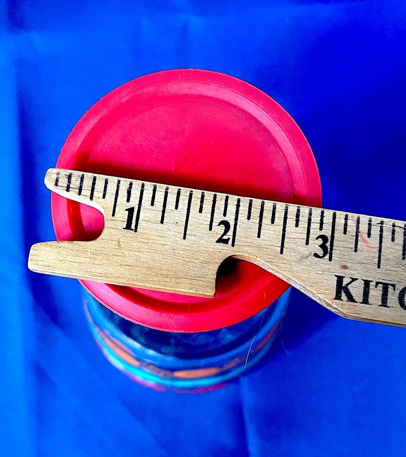 May include: A red lid covers a blue and orange container. A wooden ruler is placed across the lid, measuring approximately 3 inches. The container sits against a bright blue background.