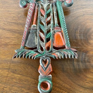 May include: A cast iron trivet with a green, orange, and white design. The trivet features three stylized brooms and a leafy design. The trivet has a heart-shaped handle with a ring at the bottom.