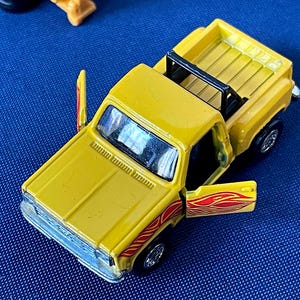 May include: A yellow toy pickup truck with a black roll bar and open doors. The truck has red and orange flame decals on the doors. The truck is a miniature replica of a classic pickup truck.