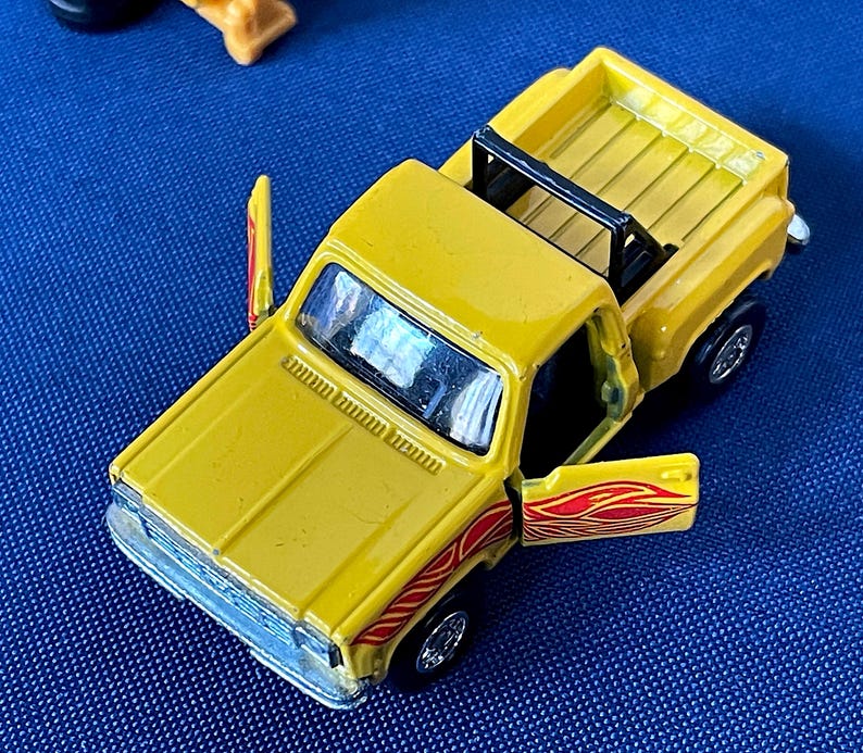 May include: A yellow toy pickup truck with a black roll bar and open doors. The truck has red and orange flame decals on the doors. The truck is a miniature replica of a classic pickup truck.