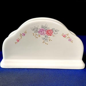 May include: A white ceramic napkin holder with a floral design featuring pink and red roses, gray leaves, and gold accents. The holder has a curved top and a rectangular base, suitable for kitchen or dining decor.