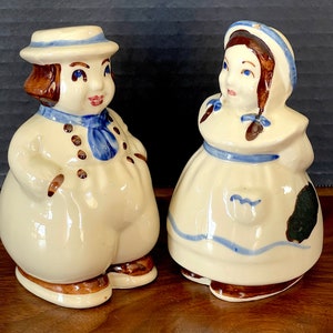 以下が含まれることがあります： 男の子と女の子の形をしたヴィンテージのセラミック製塩コショウ入れのペア。男の子は青と白の帽子と青いスカーフを着用しています。女の子は青と白のドレスと髪に青いリボンを着用しています。両方の塩コショウ入れは茶色の髪とペイントされたディテールを持っています。