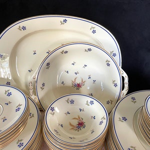 Puede incluir: Un juego de vajilla de porcelana vintage con fondo crema y estampado floral azul. El juego incluye una fuente ovalada grande, un tazón para servir y una pila de platos y platillos.