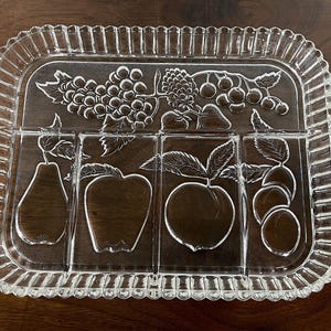 May include: Clear glass serving tray with a rectangular shape and a decorative, scalloped edge. The tray features embossed fruit designs, including grapes, pears, apples, peaches, and plums, divided into separate sections. The tray has two rounded handles.