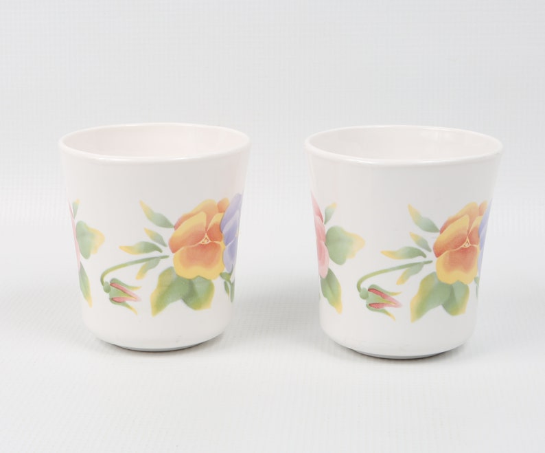 May include: Two white ceramic mugs with a floral design featuring yellow, pink, and purple pansies. The mugs have a simple, elegant design.