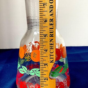 May include: A clear glass pitcher with a red lid, decorated with orange and red fruit designs and green leaves. The pitcher is approximately 8 inches tall.