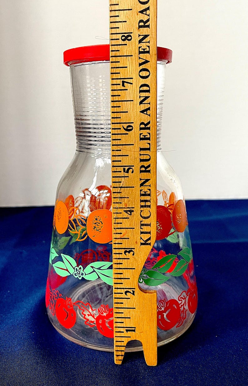 May include: A clear glass pitcher with a red lid, decorated with orange and red fruit designs and green leaves. The pitcher is approximately 8 inches tall.