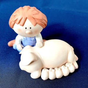 Könnte beinhalten: Keramikfigur, die ein Kind in blauen Latzhosen zeigt, das auf einem großen Schwein sitzt, mit mehreren kleinen Ferkeln darunter. Das Kind hat rotes Haar und Sommersprossen. Das Schwein und die Ferkel sind hellrosa.