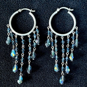 May include: A pair of silver hoop earrings with dangling strands of blue and iridescent crystal beads. The earrings have a cascading design, with the beads varying in size and shape, creating a shimmering effect. The background is a dark, textured surface.