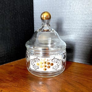 May include: A clear glass jar with a gold lid and a grape design. The jar has a white background with black outlines and gold grapes.