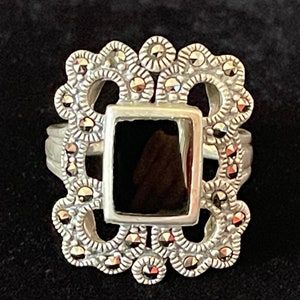 Puede incluir: Un anillo de plata con una piedra negra engastada en un diseño de filigrana. El anillo está decorado con pequeñas piedras brillantes.