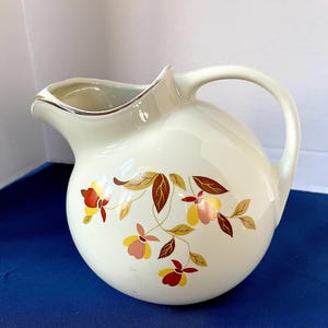 May include: Cream-colored ceramic pitcher with a rounded body and a curved handle. The pitcher features a floral design in shades of red, yellow, and brown. The rim is trimmed with a gold accent. A classic piece for serving beverages.