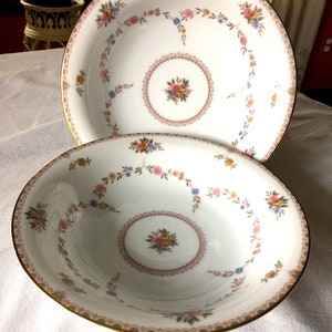 May include: Two white porcelain bowls with floral patterns. Each bowl has a central floral bouquet and a decorative border of pink, blue, and yellow flowers. The rims are trimmed with gold. The bowls are on a white surface.
