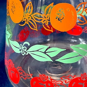 May include: Clear glass jar with a colorful fruit design. The jar features orange, red, and green fruit and floral patterns. The design includes orange circles, red tomatoes, and green leaves. The jar is likely for kitchen or home decor.