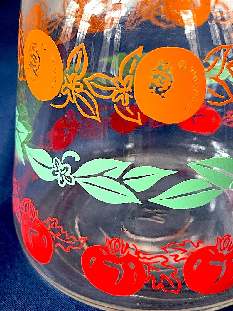 May include: Clear glass jar with a colorful fruit design. The jar features orange, red, and green fruit and floral patterns. The design includes orange circles, red tomatoes, and green leaves. The jar is likely for kitchen or home decor.