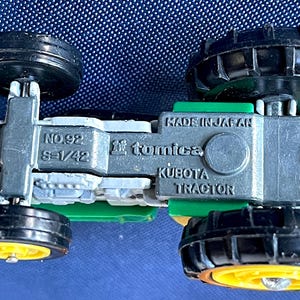 May include: A miniature Kubota tractor toy, viewed from below, with black wheels and a green and silver body. The toy is marked "Made in Japan" and "Tomica". The wheels have yellow centers.