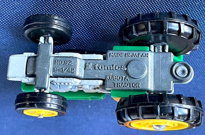 May include: A miniature Kubota tractor toy, viewed from below, with black wheels and a green and silver body. The toy is marked "Made in Japan" and "Tomica". The wheels have yellow centers.