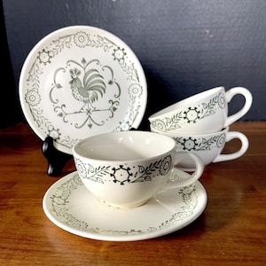 May include: A set of cream-colored ceramic teacups and saucers featuring a dark green printed pattern. The saucers display a central rooster motif surrounded by floral and leaf borders, while the cups feature a matching floral band design.