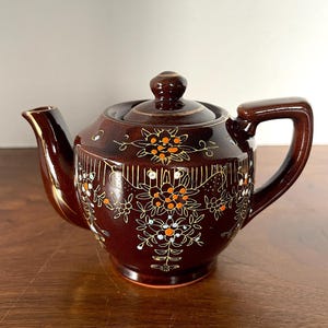 Pode incluir: Uma chaleira de cerâmica marrom com um design floral em tons de laranja, branco e dourado. A chaleira tem uma tampa e uma alça.