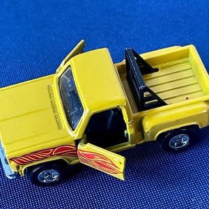 May include: A yellow toy pickup truck with the doors open, revealing a black interior. The truck has red and orange flame decals on the side and a black roll bar in the bed. The wheels are black with silver rims.