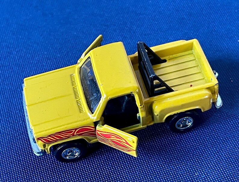 May include: A yellow toy pickup truck with the doors open, revealing a black interior. The truck has red and orange flame decals on the side and a black roll bar in the bed. The wheels are black with silver rims.