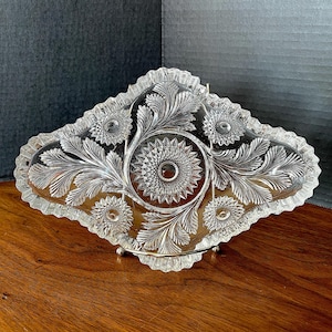 Puede incluir: Un plato de servir de vidrio transparente con un borde festoneado y un diseño floral. El plato tiene un patrón de diamante y está decorado con hojas y flores.