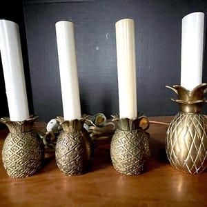 May include: A set of four brass pineapple-shaped candle lamps, each featuring a textured metallic base and a tall, cylindrical white candle sleeve. The lamps include electrical cords and plugs, with three showing a detailed cross-hatch pattern.