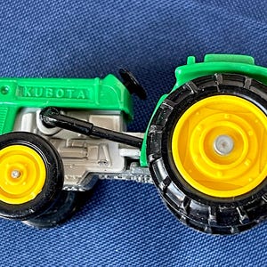 May include: A green and silver toy tractor with the word "KUBOTA" printed on the hood. The tractor has black tires with yellow rims. The tractor is a miniature replica of a farm vehicle.