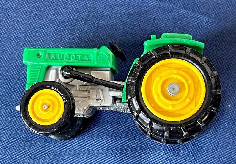 May include: A green and silver toy tractor with the word "KUBOTA" printed on the hood. The tractor has black tires with yellow rims. The tractor is a miniature replica of a farm vehicle.