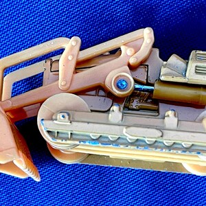 May include: A tan and beige toy excavator with a blue gem detail. The toy has a scoop, a cab, and tracks. The excavator is made of plastic and has some blue paint details.