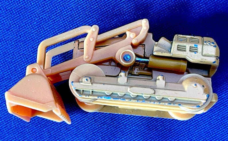 May include: A tan and beige toy excavator with a blue gem detail. The toy has a scoop, a cab, and tracks. The excavator is made of plastic and has some blue paint details.