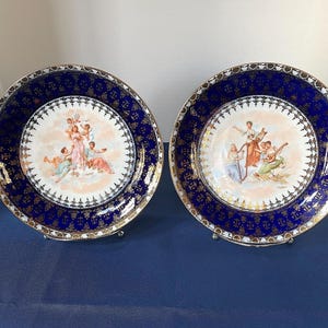 May include: Two decorative plates with a blue and gold rim. The center of each plate features a detailed illustration of figures in a classical style. The plates are displayed on silver stands against a blue background. The plates are likely for decorative purposes.