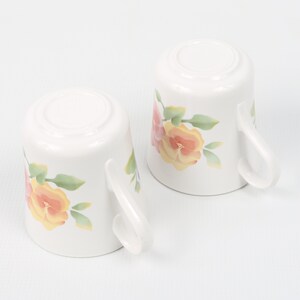 May include: Two white ceramic mugs with floral designs. The mugs feature pink, yellow, and green flowers.