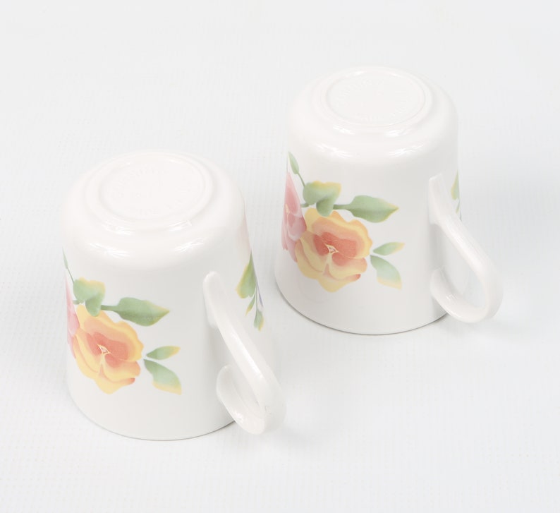 May include: Two white ceramic mugs with floral designs. The mugs feature pink, yellow, and green flowers.