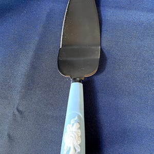 Puede incluir: Una paleta para tartas con una hoja de acero inoxidable y un mango azul claro. El mango presenta un diseño en relieve blanco. La paleta mide unos 25 cm de largo.