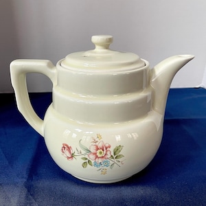 May include: Cream-colored ceramic teapot with a lid and a curved handle. The teapot features a floral design with pink, red, and blue flowers. The spout is curved, and the teapot is set against a blue background.