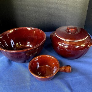 May include: A set of three glazed ceramic kitchenware pieces in a rich, reddish-brown color. The set includes a large bowl, a lidded casserole dish, and a small handled bowl. The pieces are arranged on a blue surface.