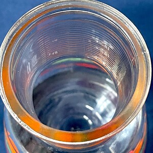 May include: A clear glass jar with a wide opening and a slightly orange-tinted rim. The jar has a cylindrical shape with vertical ridges on the inside. The bottom of the jar has colorful designs.
