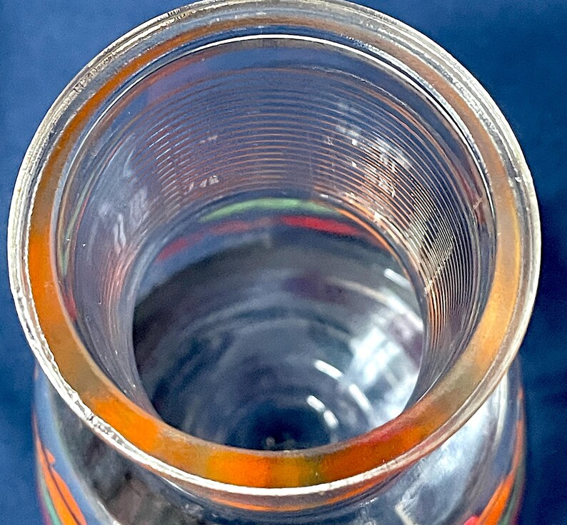 May include: A clear glass jar with a wide opening and a slightly orange-tinted rim. The jar has a cylindrical shape with vertical ridges on the inside. The bottom of the jar has colorful designs.