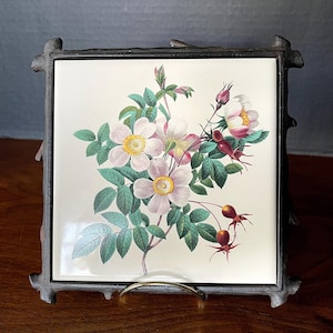 May include: A vintage ceramic tile with a floral design featuring pink and white roses with green leaves and red berries. The tile is framed in a black metal frame with a decorative twig design.