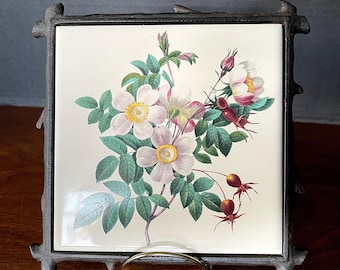 May include: A vintage ceramic tile with a floral design featuring pink and white roses with green leaves and red berries. The tile is framed in a black metal frame with a decorative twig design.