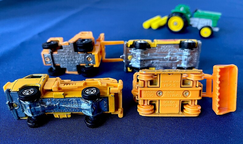 May include: A collection of toy construction vehicles, including a yellow dump truck, a bulldozer, and a green tractor. The toys are made of metal and plastic, with black wheels. The vehicles are arranged on a blue surface.