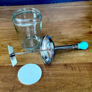Puede incluir: Un frasco de vidrio transparente con una marca de capacidad de 18 oz, un conjunto de tapa metálica con una varilla central y un resorte, un disco circular blanco plano y un mango de pera de color turquesa brillante en la varilla.