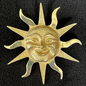 May include: A gold-toned sun brooch with a smiling face and a textured surface. The sun has 12 rays.