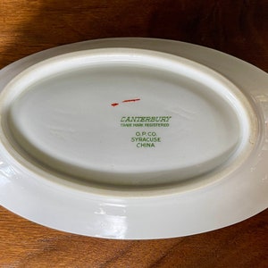 May include: White oval ceramic dish with a green and red inscription that reads "Canterbury Trade Mark Registered O.P. Co. Syracuse China".