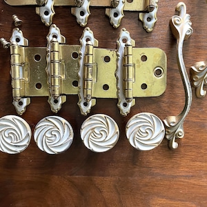 May include: A collection of vintage brass hardware, including hinges, drawer pulls, and a handle. The hardware features white accents and decorative detailing. Screws are included.