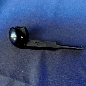 May include: A black pipe with a rounded bowl and a long, flat stem. The pipe has text on the stem. The pipe is set against a blue background.