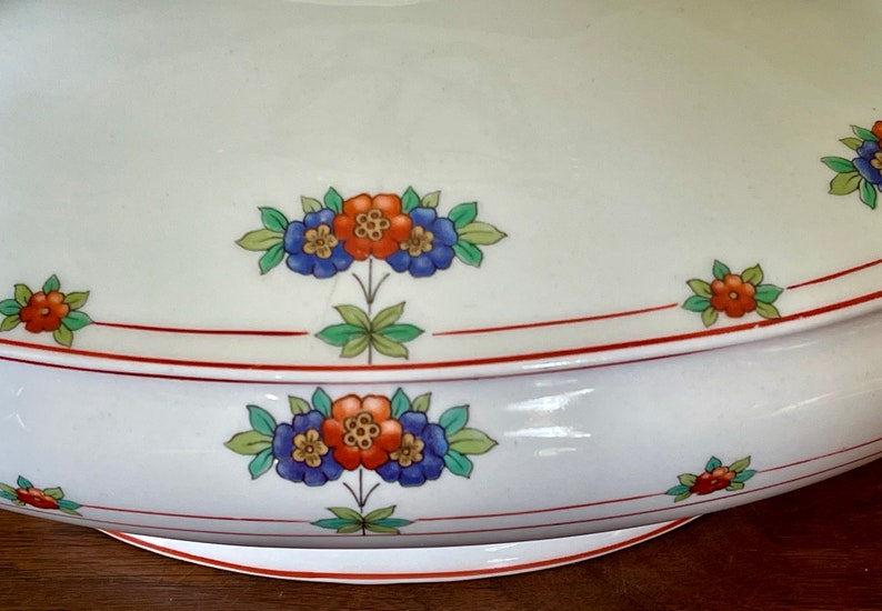 May include: White ceramic serving dish with a red and orange floral design. The dish has a scalloped edge and a red rim.