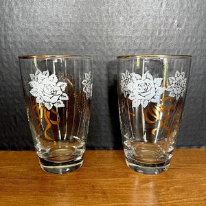 Vasos altos LIBBEY (2 unidades). Se venden juntos. Mediados del siglo XX. Rosas blancas, lazos dorados, puntos y ribetes sobre cristal transparente. 13,3 cm de alto. Sin desgaste ni daños.